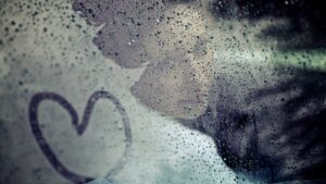 Hochzeitsfoto Idee im Regen: Die Braut hinter einer verregneten Autoscheibe, die von innen leicht beschlagen ist, und auf die mit dem Finger ein Herz gemalt wurde